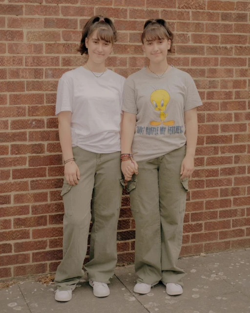 Two young women holding hands pose in front of a brick wall, wearing casual T-shirts, olive cargo pants, and white sneakers, medium shot.