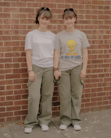 Two young women holding hands pose in front of a brick wall, wearing casual T-shirts, olive cargo pants, and white sneakers, medium shot.