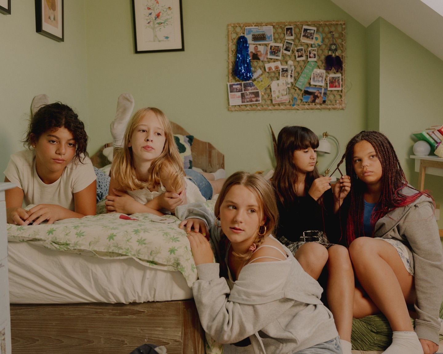 Group of five young girls in casual clothing indoors: one lying on bed, two braiding hair, and two seated, with neutral expressions.