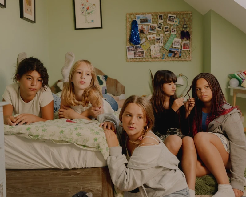 Group of five young girls in casual clothing indoors: one lying on bed, two braiding hair, and two seated, with neutral expressions.