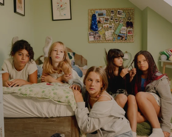 Group of five young girls in casual clothing indoors: one lying on bed, two braiding hair, and two seated, with neutral expressions.