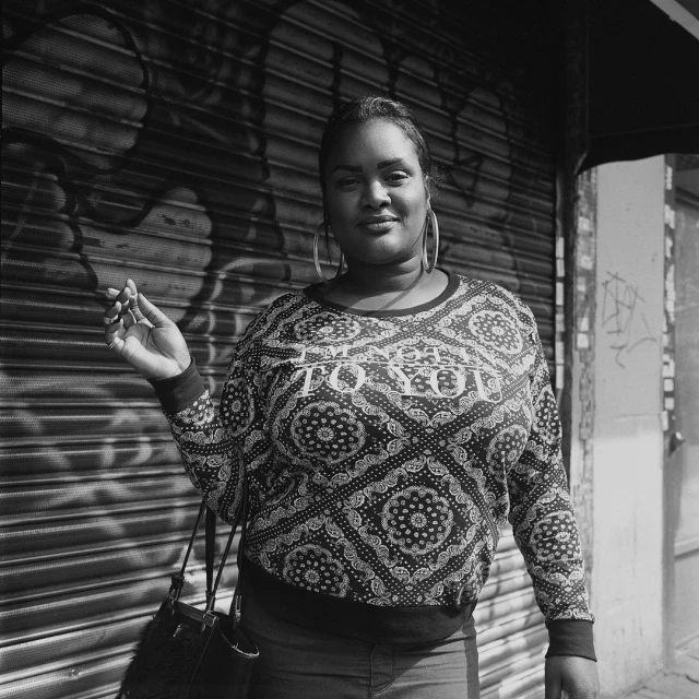 Black and white portrait of a woman with large hoop earrings, wearing a patterned shirt and holding a cigarette, standing in front of a closed roller door.