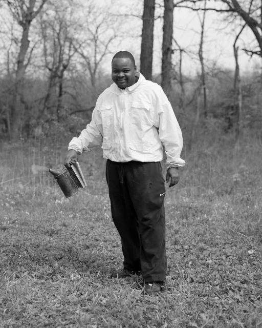 Monochrome portrait of an adult man standing in a grassy field, holding a beekeeping smoker, with bare trees in the background.