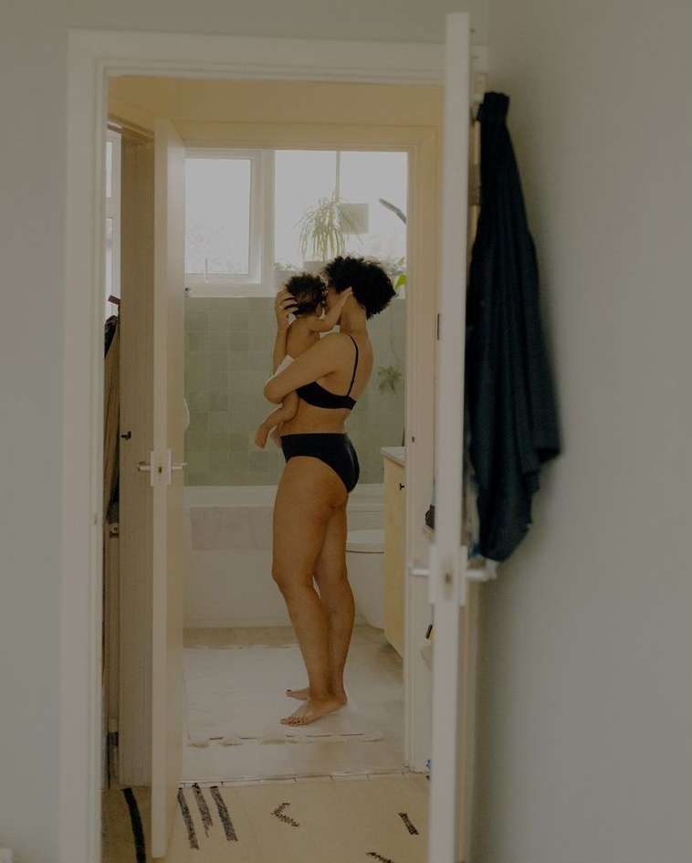 A woman in a black bra and underwear holds a baby in an indoor setting, viewed through a doorway. The woman stands on a patterned rug.
