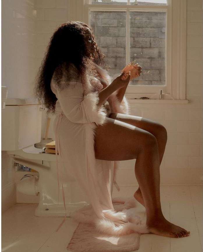 An adult woman with long dark curly hair sits in a bathroom in a light pink robe with decorative fur trim holding a sponge.
