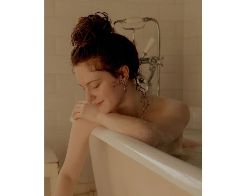 A serene portrait of a woman with red hair relaxing in a vintage-style bathtub filled with water, showcasing soft, natural light.