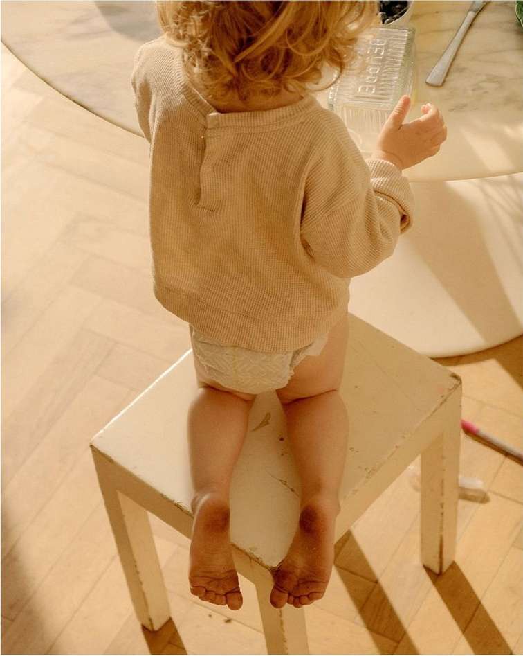 Toddler seated on a white stool, facing away from the camera, wearing a beige sweater, with dirty feet. Indoors on a light floor.