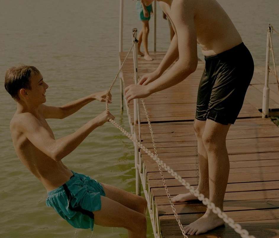 Two teenage boys on a wooden dock are holding onto a chain, preparing to jump into the water on a sunny day with a happy expression.