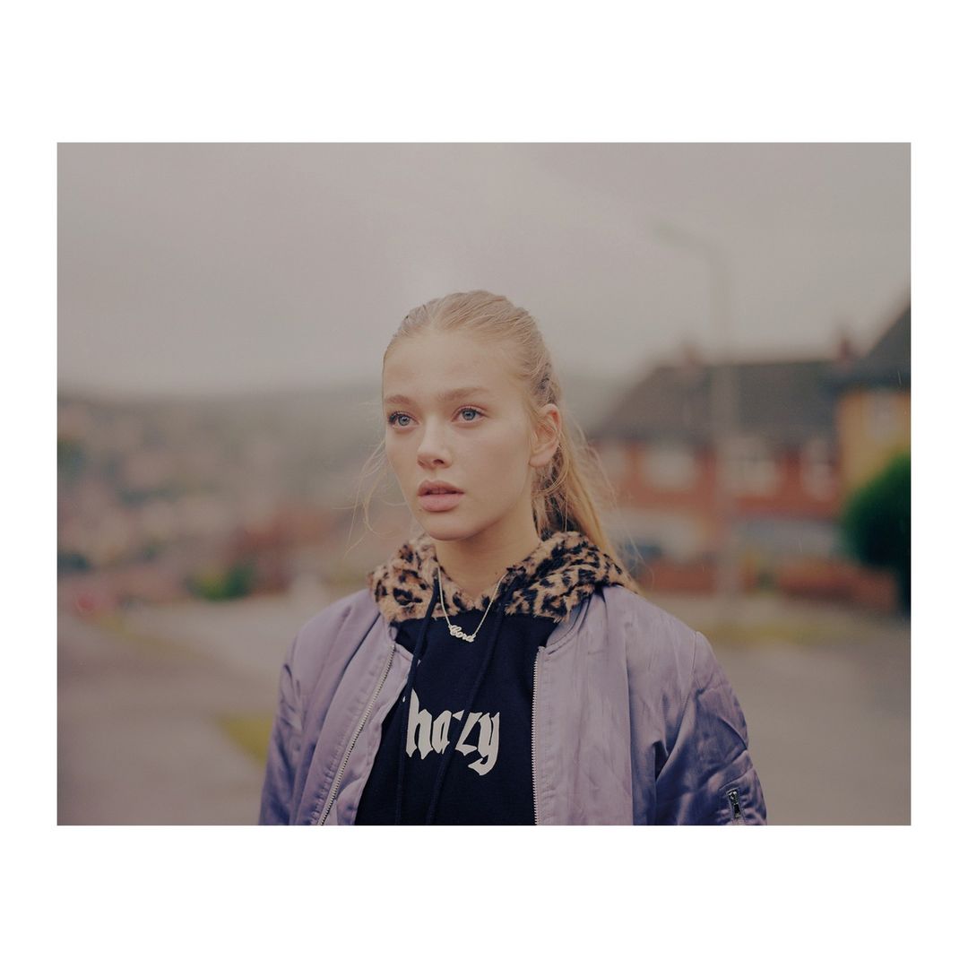 Blonde woman in a hoodie and bomber jacket, stands outdoors on a city street on an overcast day, houses in the background.