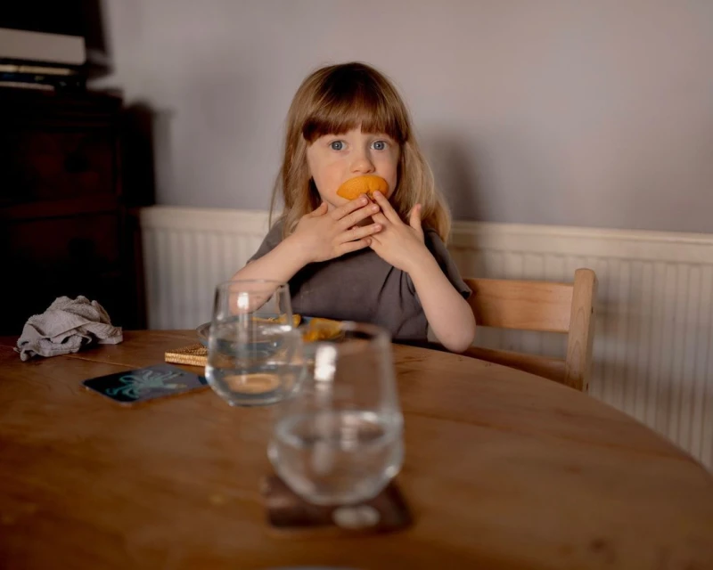 A young child sits at a table with glasses of water and puts a slice of orange in their mouth indoors, medium shot.