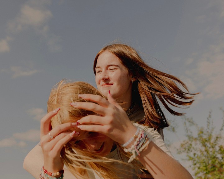 Two young women embracing outdoors under a sunny sky, with light wind blowing through their hair, one giving the other a piggyback ride.