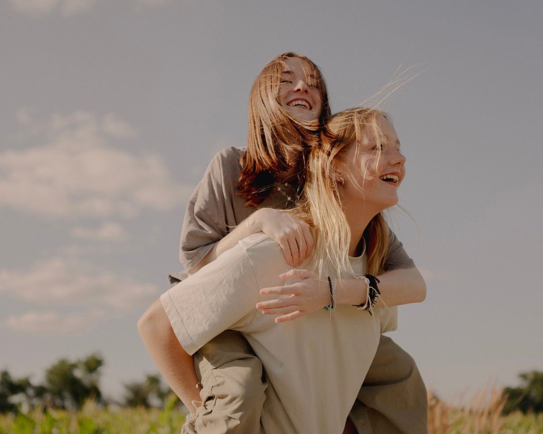 Two happy young women in casual clothing are smiling in a field with a blue sky and scattered clouds on a sunny day.