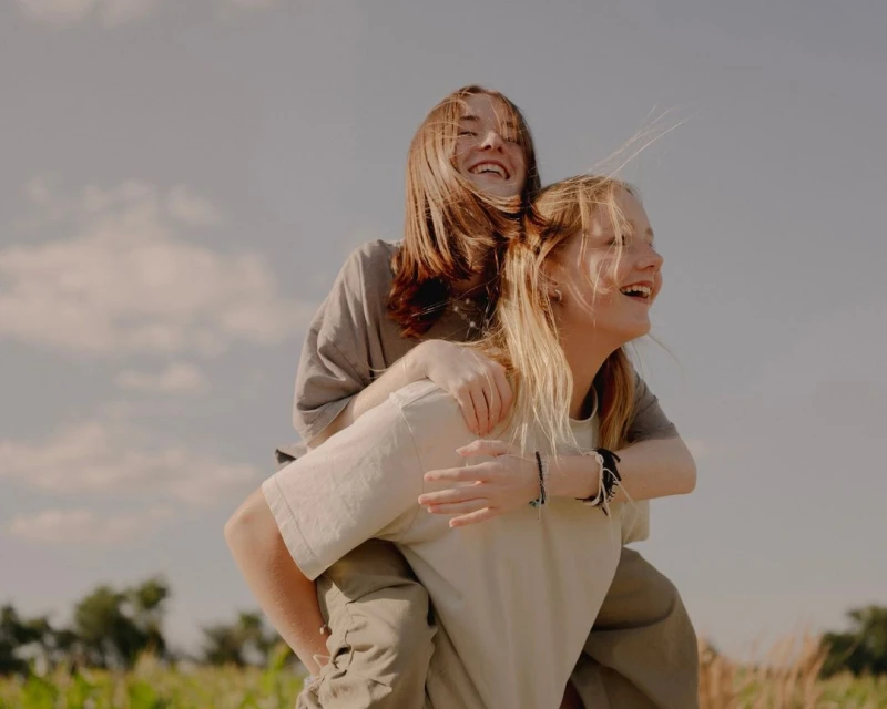 Two happy young women in casual clothing are smiling in a field with a blue sky and scattered clouds on a sunny day.