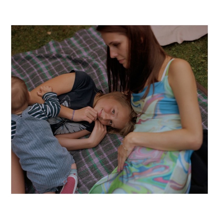 Family portrait outdoors: Mother and two children relaxing on a plaid blanket in a grassy area, enjoying a quiet moment.