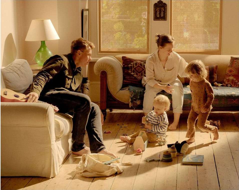 Indoor family portrait captures father, mother, toddler, and baby son relaxing on living room furniture with toys and natural light.