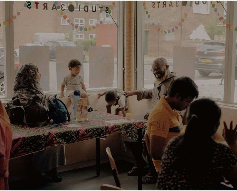 Group of diverse adults and children gather indoors with large window, colorful decorations, and table with objects.