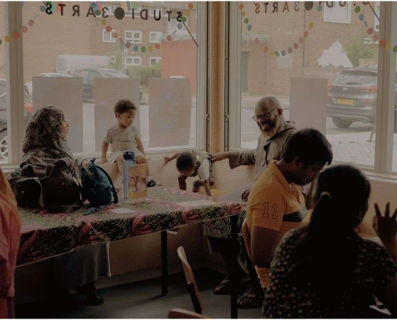 Group of diverse adults and children gather indoors with large window, colorful decorations, and table with objects.