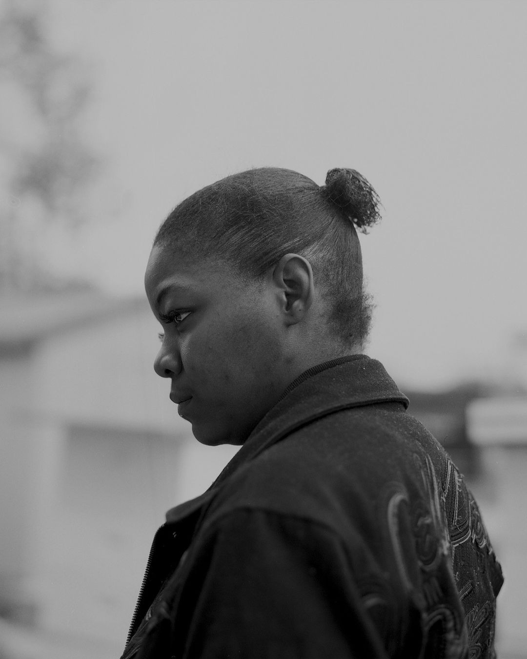 Portrait of a woman wearing a dark jacket with embroidered details, looking away, hair in a bun, against a blurred background.