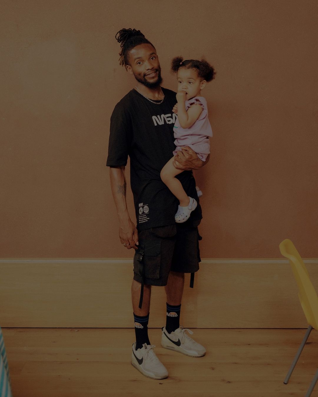 A man holding a toddler in an indoor studio setting. The man wears a NASA t-shirt, socks with clouds, and white shoes. The child wears white shoes.