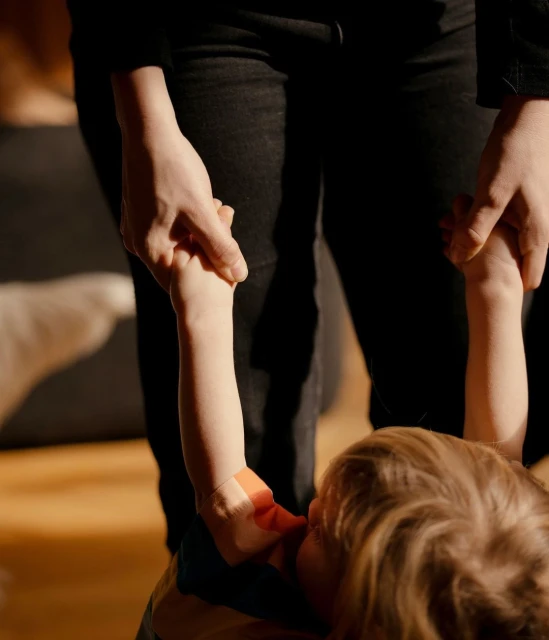 A parent holds the hands of a toddler, who is also holding the parent's hand, in a home interior with wooden floors and warm lighting.