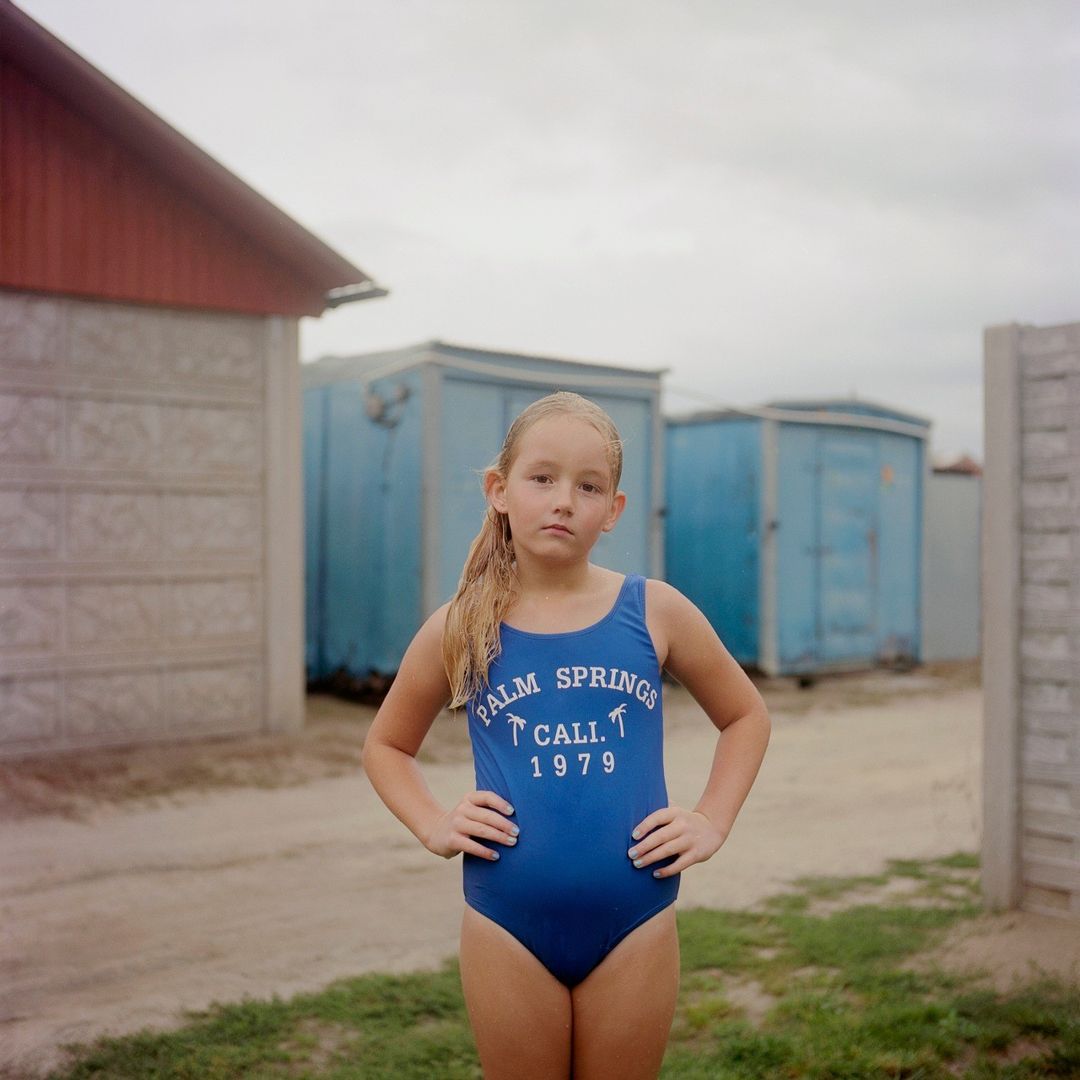 Young girl in a blue one-piece swimsuit posing with hands on her hips outdoors in front of small structures by Tori Ferenc