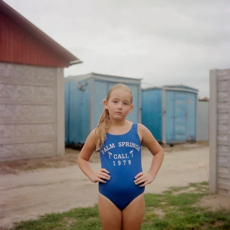 Young girl in a blue one-piece swimsuit posing with hands on her hips outdoors in front of small structures by Tori Ferenc