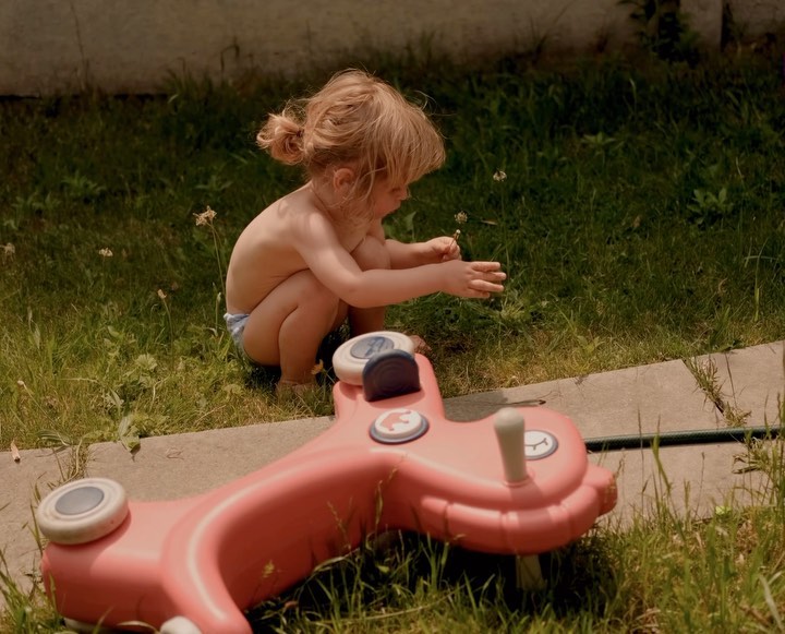 A toddler sits on green grass next to a pink toy car, wearing a diaper and looking at small white flowers in a summery outdoor scene.