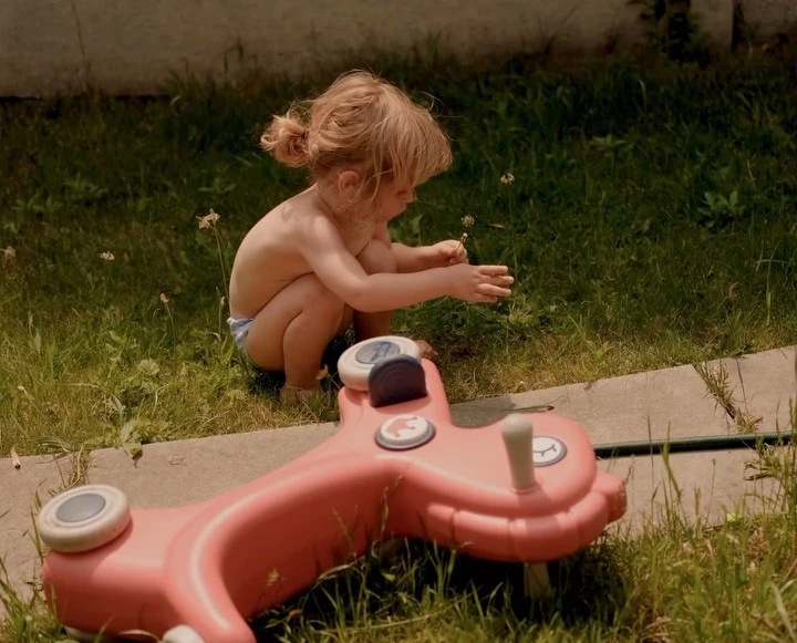 A toddler sits on green grass next to a pink toy car, wearing a diaper and looking at small white flowers in a summery outdoor scene.