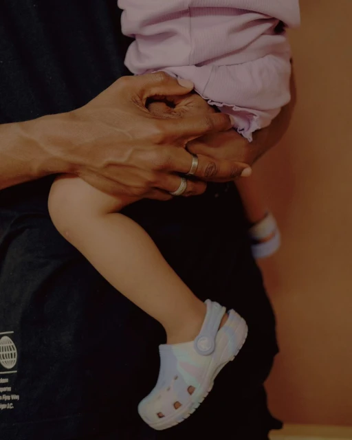 Close-up of a child being held, showcasing their pastel-colored Crocs shoes, with an adult in a black t-shirt partially visible.