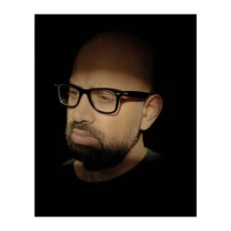 Portrait of a bald man wearing dark-rimmed glasses against a dark background, photographed by Tori Ferenc