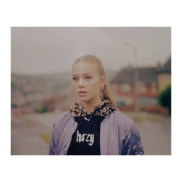 Portrait of a woman wearing a lavender bomber jacket over a black hooded top outdoors in London, photographed by Tori Ferenc.