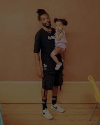 Black man holding a young child in a pink top, standing indoors against a warm brown wall photographed by Tori Ferenc.