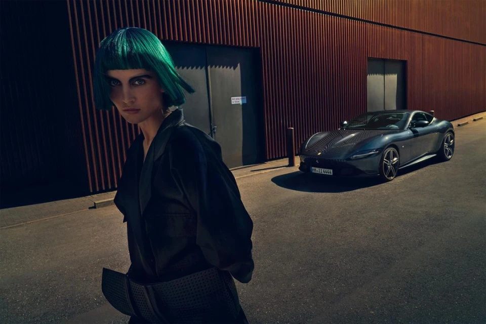 Stylish woman with green hair poses next to a modern grey Ferrari Roma coupe on a city street with a rust-colored wall backdrop.
