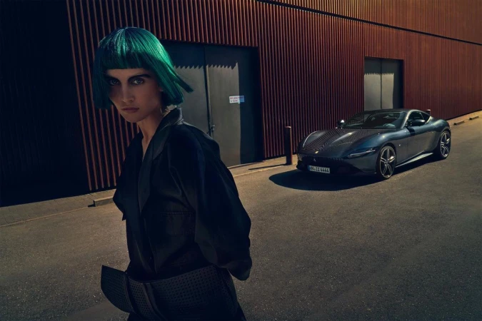 Stylish woman with green hair poses next to a modern grey Ferrari Roma coupe on a city street with a rust-colored wall backdrop.
