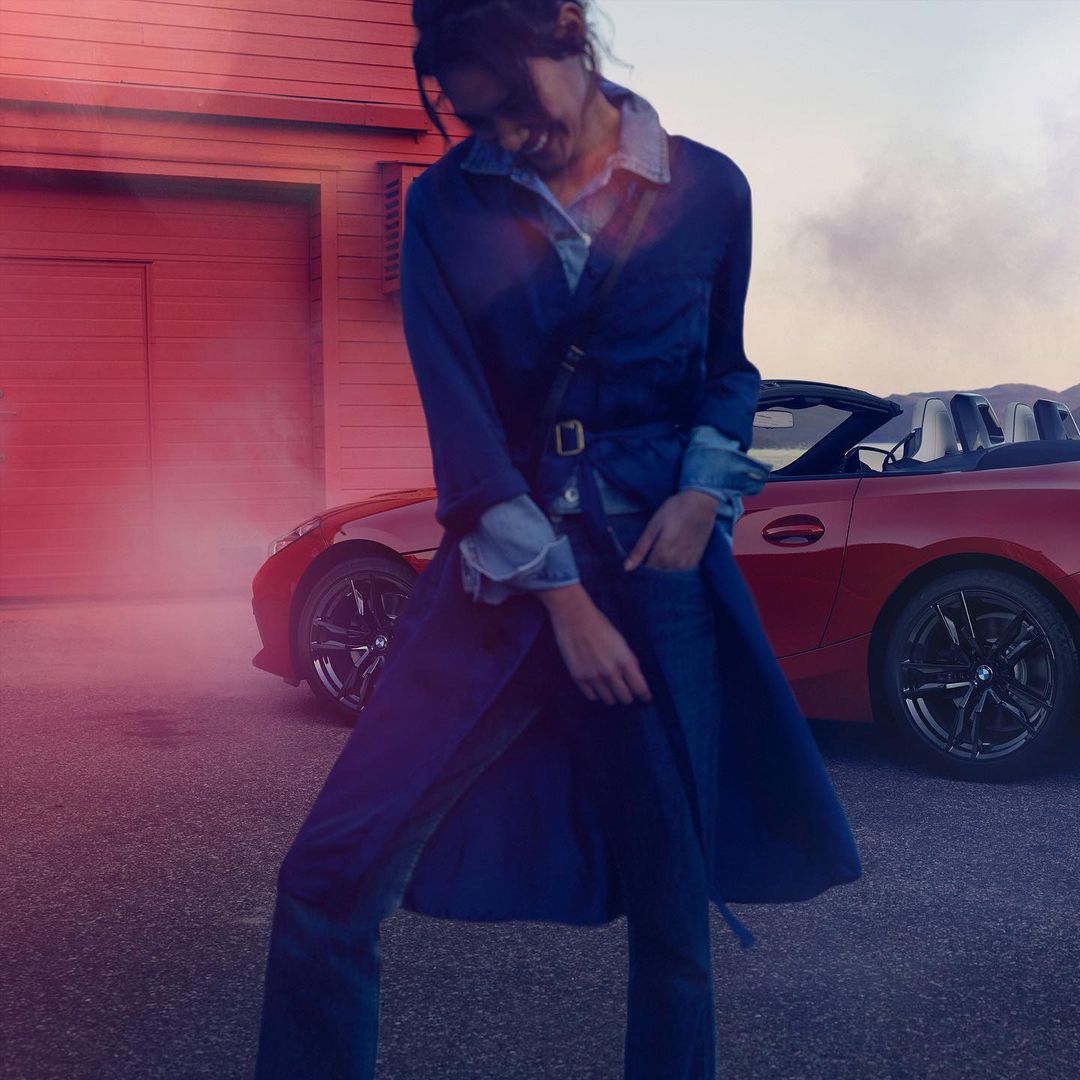 Adult woman in denim trench coat poses next to a red BMW convertible roadster in an outdoor setting with a building in the background.