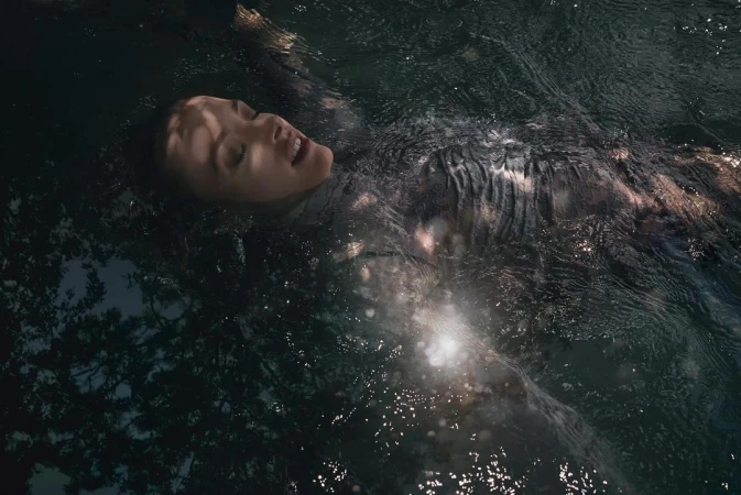 Woman floating in dark water with sunlit ripples across her body, captured from an overhead angle by Torsten Klinkow.