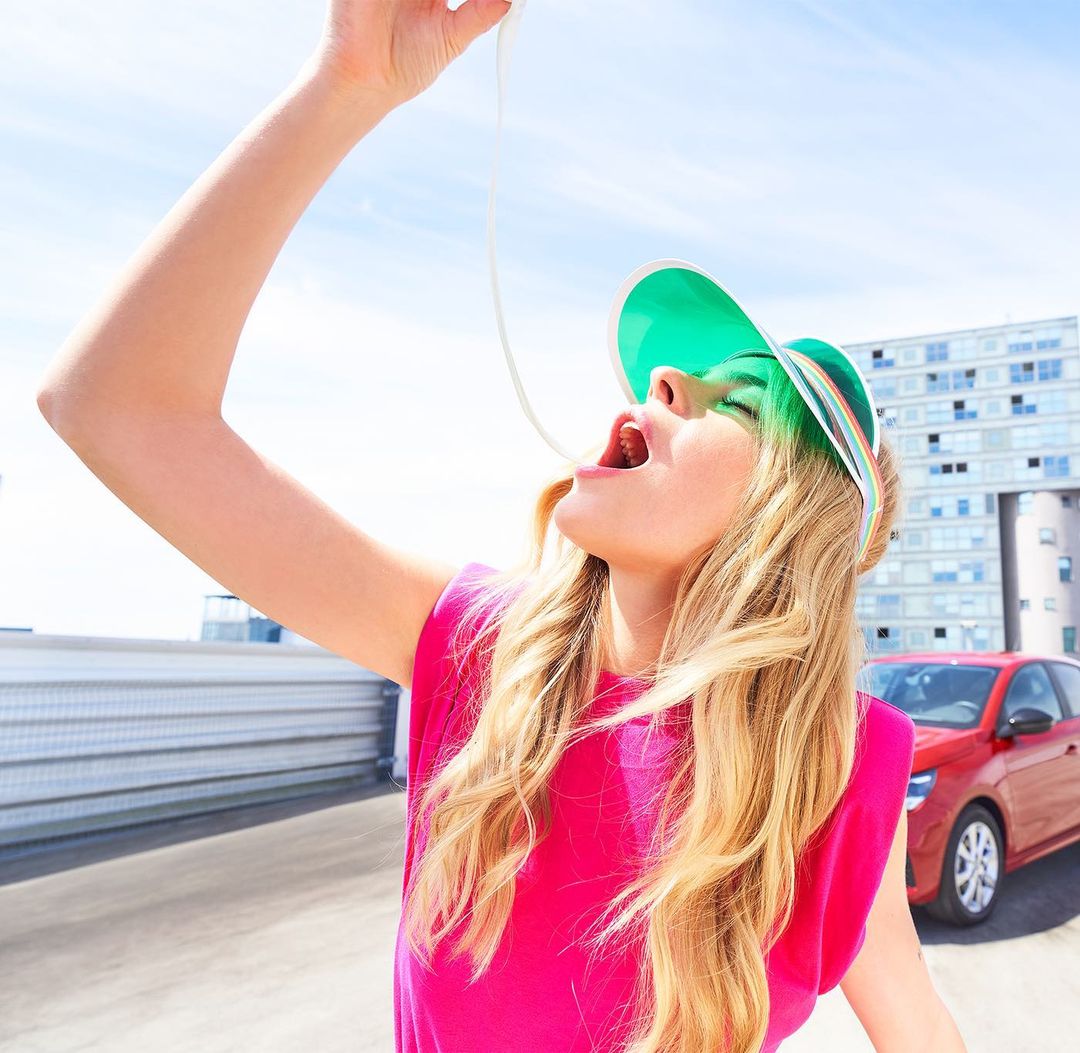 A young adult woman in a pink tank top eating a white string substance with a car and modern building in the background on a sunny day.