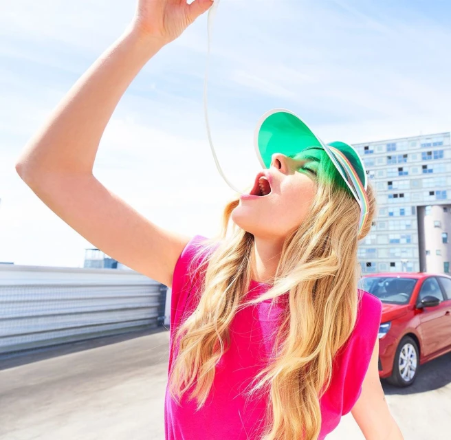 A young adult woman in a pink tank top eating a white string substance with a car and modern building in the background on a sunny day.