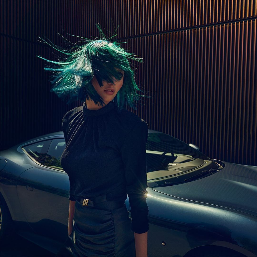 A woman with blue-green hair poses near a dark blue sports car, set against a vertically-striped dark brown wall on a sunny day.