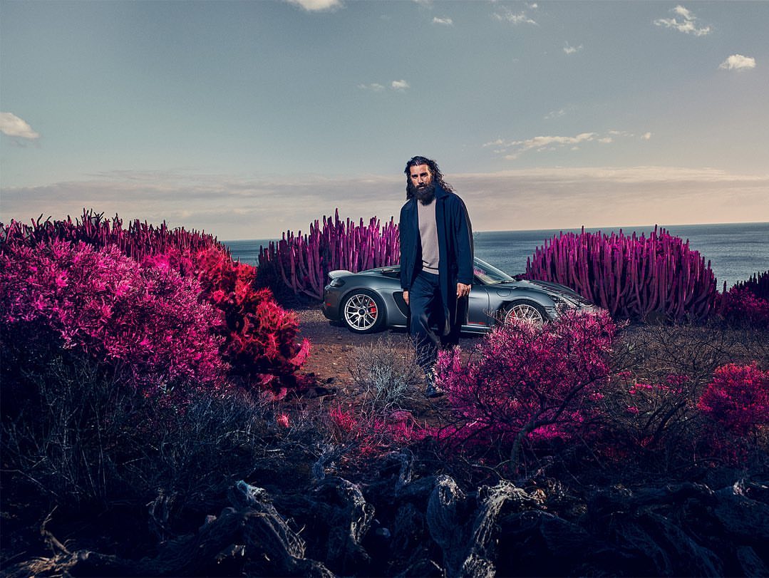 Man in coat standing next to a grey Porsche car on a desert landscape with magenta-colored flora and the ocean in the background.