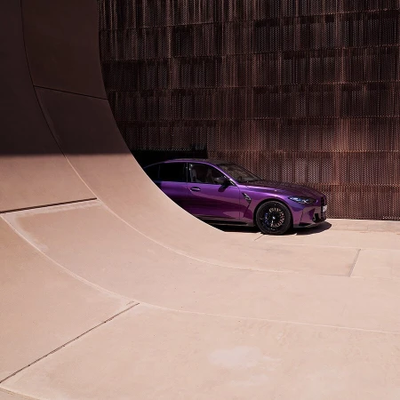 A purple BMW sedan is parked next to a contemporary architecture with a light brown exterior wall and a perforated wall alongside.