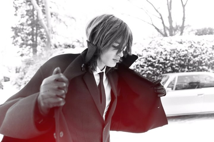 Stylish man in suit and gloves opens his coat outdoors in a wintery setting, with a white car visible in the background.