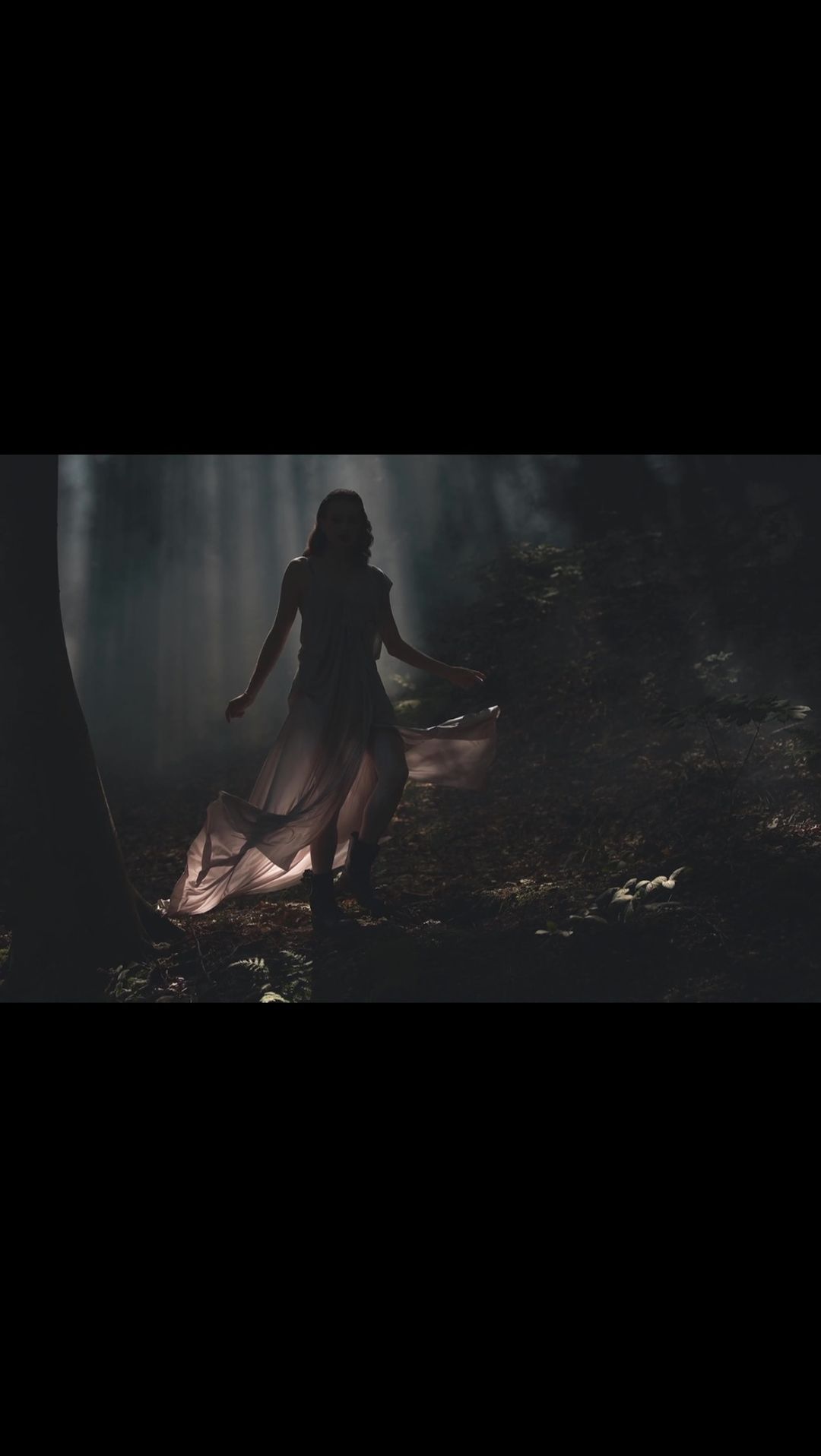 Woman in flowing dress walks in a dark forest. Sunlight shines through the trees, creating a fantasy or ethereal atmosphere.