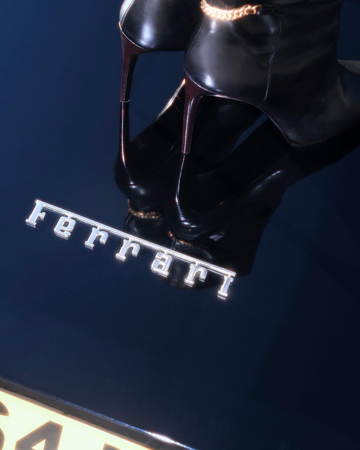 High-heeled black boots with a gold chain accent placed on a glossy surface with a chrome Ferrari logo and car license plate.