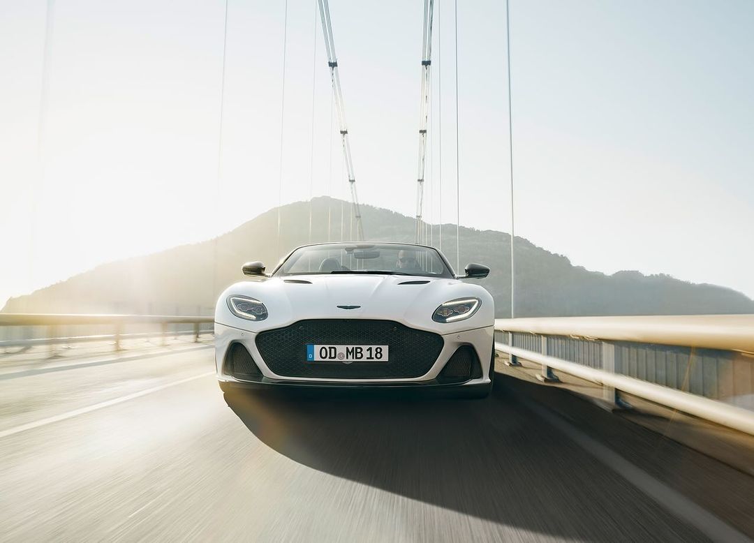 White Aston Martin convertible drives on a bridge, with a hill in the background on a sunny day, capturing automotive elegance.