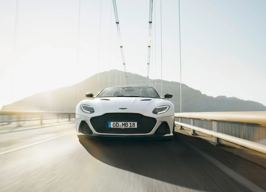 White Aston Martin convertible drives on a bridge, with a hill in the background on a sunny day, capturing automotive elegance.