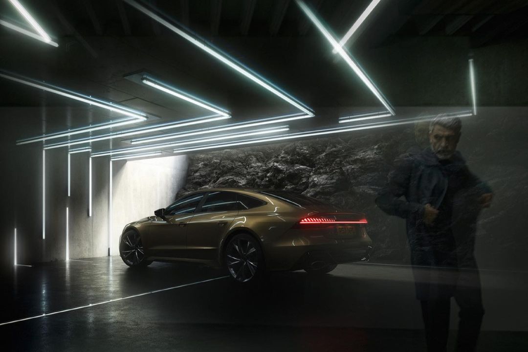 A gold Audi car is parked indoors under neon lights next to a man in a blue coat standing in a dark area with a cave-like wall.