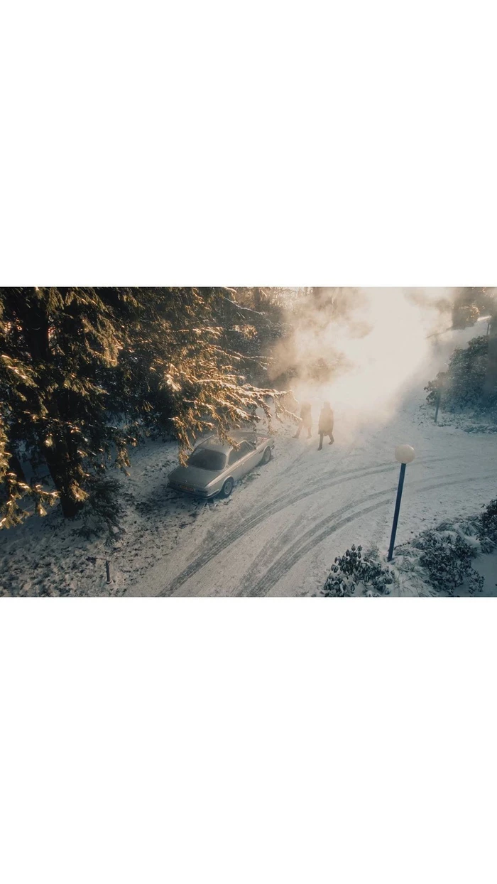 High-angle view of two people walking on a snowy path near a parked silver car and tall evergreen trees.