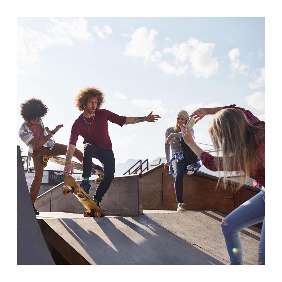 Group of young adults skateboarding at a skate park. One man on a skateboard, others watching and taking photos on a sunny day.
