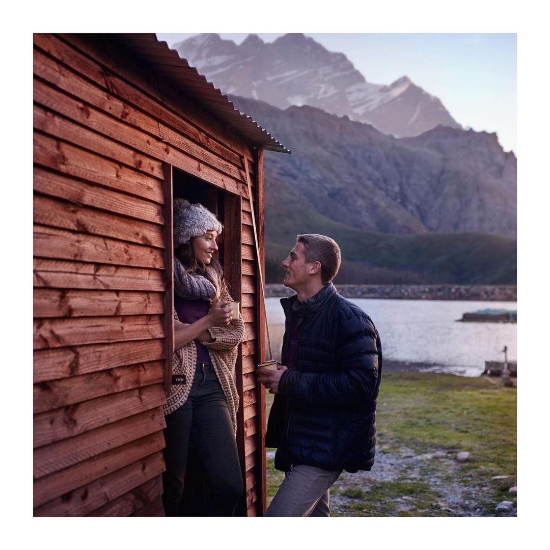 A couple stands outside a rustic wooden cabin with snowy mountains in the background, enjoying a scenic view together in natural light.